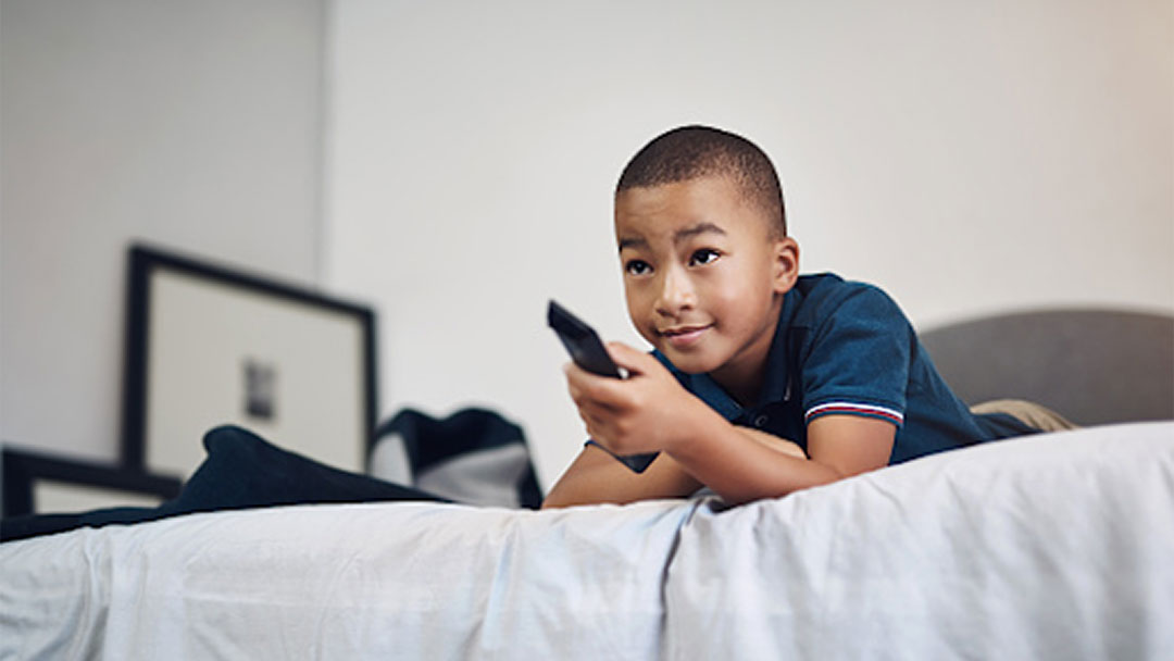 Boy laying on the bed using a remote to change a television channel
