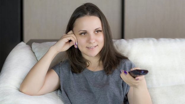 Woman sitting on white couch holding tv remote towards screen