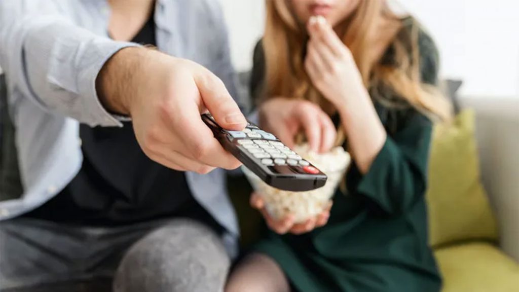 A couple sitting on a couch eating popcorn and holding a TV remote.