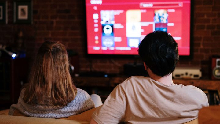 Back view of a couple sitting on a couch in a living room and watching TV.