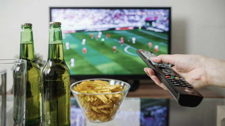 A man holding a TV remote near a bowl of potato chips and two beer bottles with a TV showing soccer in the background.