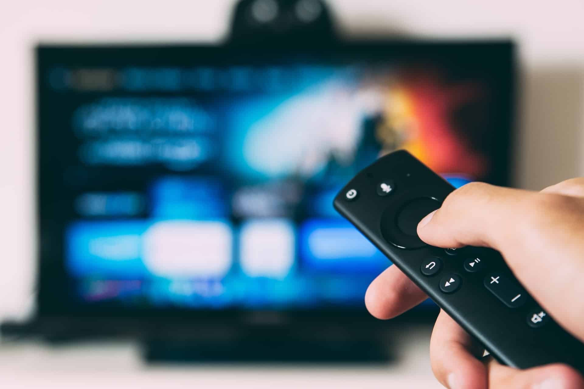 Hand holding a TV remote with a blurry TV in the background
