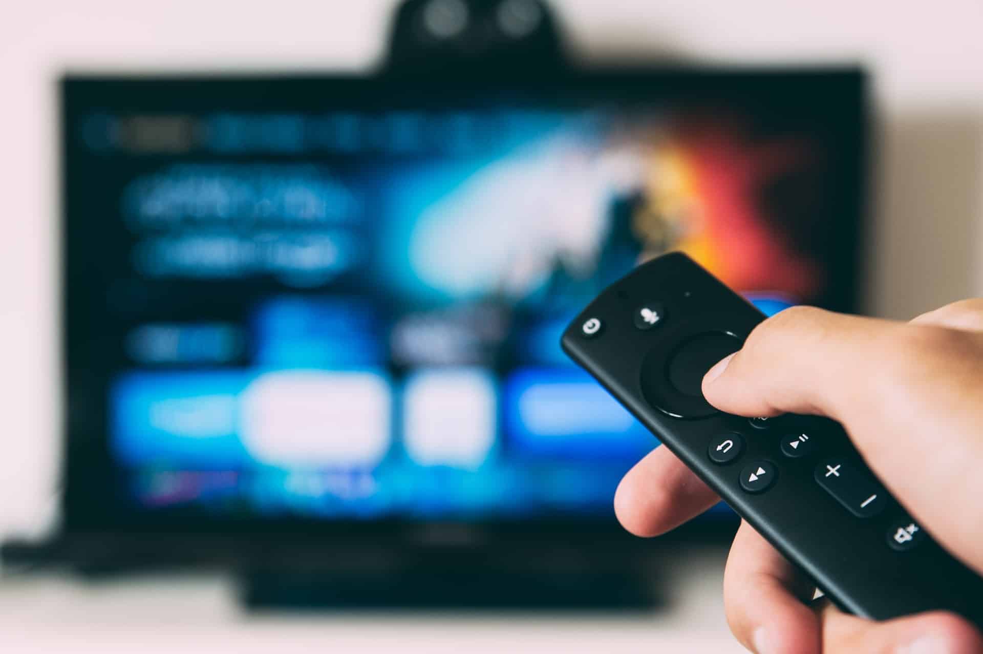 Hand holding a TV remote with a blurry TV in the background